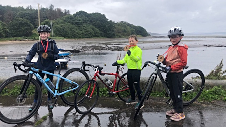 Three young boys stood with their bikes holding medals around their necks at the Scottish seaside on a cloudy day