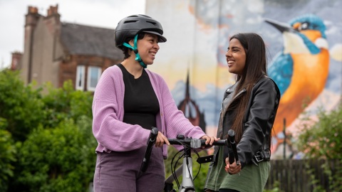 One woman in a helmet standing with a bike and another walking