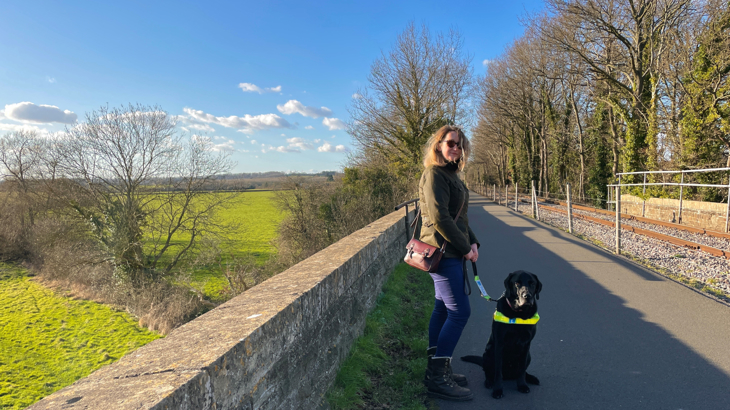 A woman called Sarah poses for a photograph with her black Labrador guide dog called Webbly. Webbly's wearing a guiding harness which Sarah's holding. The pair are stood on a former railway bridge on the Bristol Bath Railway Path, National Route 4 on a sunny spring day, overlooking the River Avon.