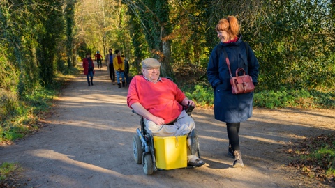 A man in a wheelchair smiling and laughing alongside a woman walking on the Festival Way in Bristol