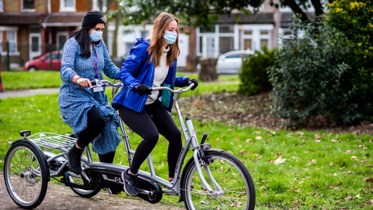 Two women wearing face masks looking happy while pedalling an adapted cycle through a park in Hounslow