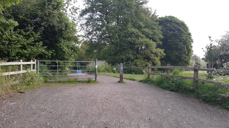 The wide entrance to a traffic-free path has a metal gate across half of it, the other half is open with a wooden post at the centre of the gap. National Cycle Network signs are on both the gate and the post. The path beyond is tree lined, some larger tress are in the distance and green shrubs are in the foreground.  The scene is lush and green with plants in full leaf.