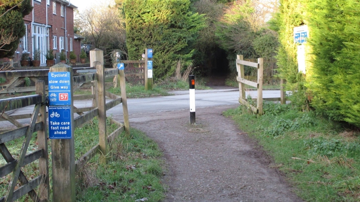 A view of a traffic-free path crossed by a narrow road. In the foreground the path has signs on a wooden fence line telling cyclists to 'slow down, give way' and 'take care fast road ahead'. As the path meets the road there is a reflective bollard in the centre. On the other side of the road the path continues along a dark tree-lined passage.