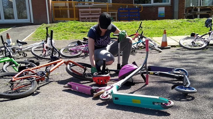 A young volunteer kneeling and looking down while fixing a bike on the ground. She is in a school playground surrounded by bikes and scooters
