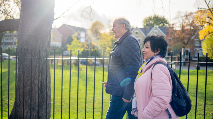 An older man and woman walk side by side smiling next to a fenced off green space in a residential area