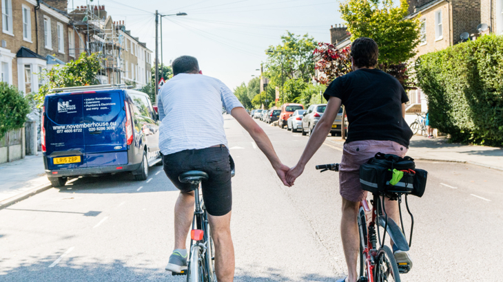 A couple are cycling along a quiet, deserted residential street on a London Quietway route. They both have short hair, are holding hands and wearing shorts and t-shirt on a bright, summery day. On the street, the houses are set back a little from the road, there are some parked cars and hedges.