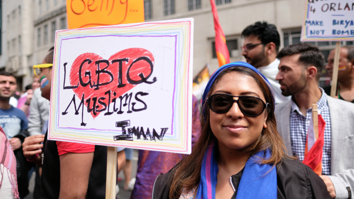 One of several Muslim participators in London's 2016 LGBT Pride Parade. A smiling woman wearing a blue and pink headscarf holds a handwritten placard which says 'LGBTQ Muslims IMAAN' behind the words is a drawn red heart and around them a rainbow border. Behind the woman are many more people in the crowd and partly obscured banners.