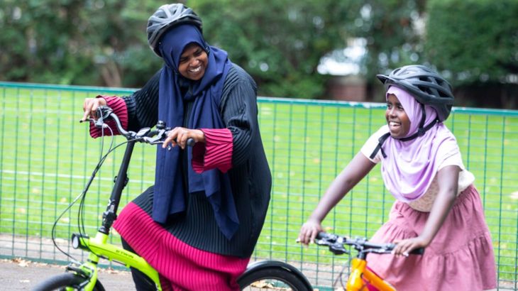 Mother and child learning how to ride a bike in a playground. In the background there is a grassed area.