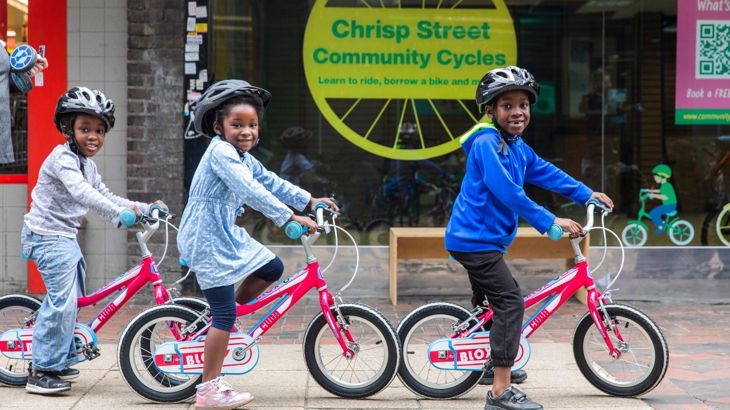 Three smiling small children on bikes, in a row, looking at the camera. They are outside the Chrisp Street Community Cycle hub in Tower Hamlets.
