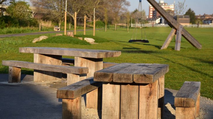 Oak park furniture - park benches and picnic tables dappled in sunlight. In the background is a lawn and the trees in autumn with no foliage.
