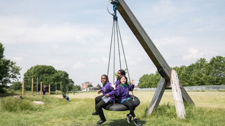 children playing on a round group swing. The swing mechanism is made of oak. The swing is made of rubber and netting. The background are trees and a lawn. It's a summer's day.