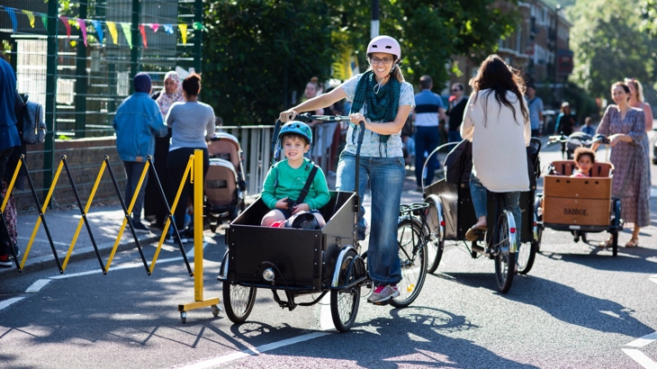 A street outside a school is closed to motor traffic and filled with pedestrians and cyclists. At the front are a woman and child in a cargo bike. The people are smiling and the sun is shining. 