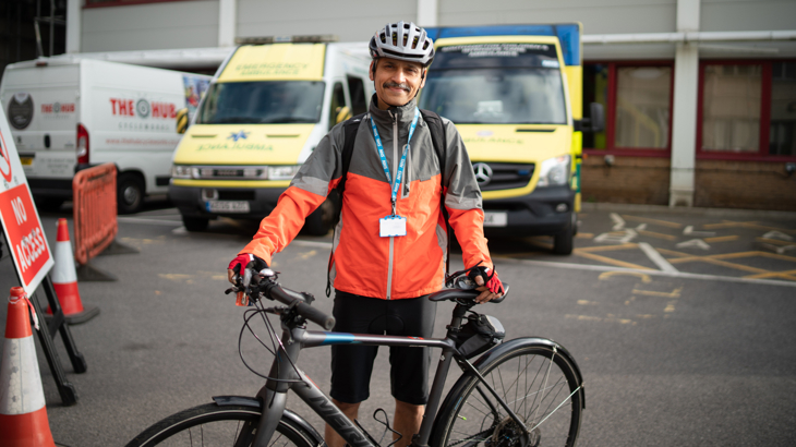 Sanjay stands holding his new grey bike on tarmac outside Southampton Hospital. He wears a helmet, waterproof jacket, cycling shorts and work lanyard. Behind him are two ambulances and hospital buildings. Sanjay is smiling and the day is dry and bright.