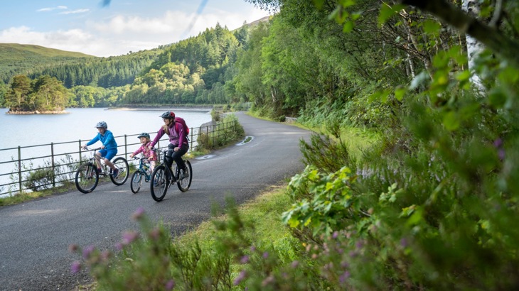 A woman and two children cycle along a path next to a loch on the Lochs and Glens Way