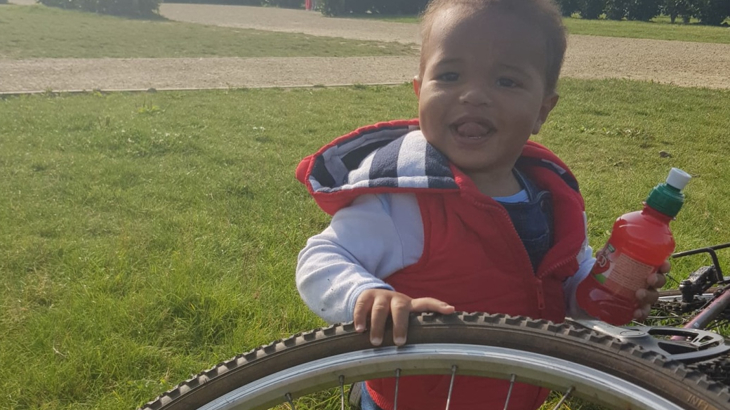 A young boy smiling at the camera sat next to and holding a stationary bike wheel on a grassy field