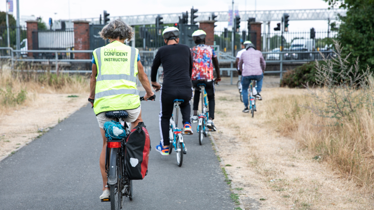 The view behind five people cycling on a traffic-free path. Four are on Brompton bikes, the other is on a standard bike with pannier. This person is wearing a hi-vis vest which says 'Cycle Confident Instructor'.  Dry summer grass is either side of the sealed surface path and the group are approaching a road junction with traffic lights and a built up urban area beyond.