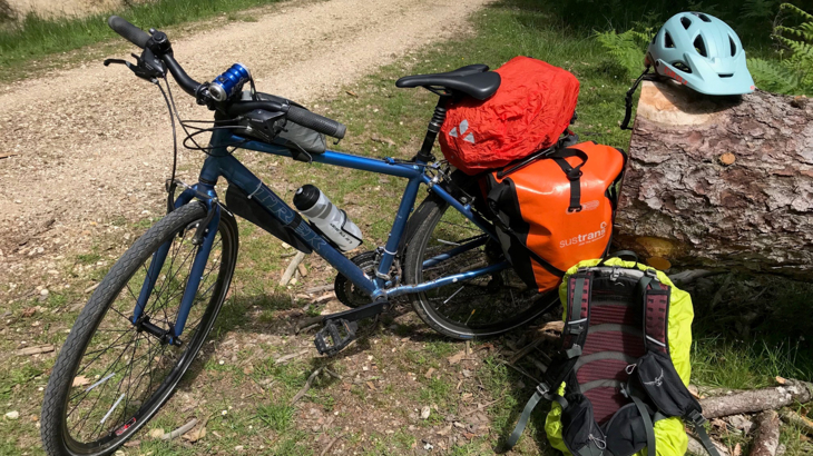 Natalie's hybrid bike resting against a log, the pannier rack has a red bag on top and two orange panniers to the side. On the ground beside the bike is a rucksack and helmet.