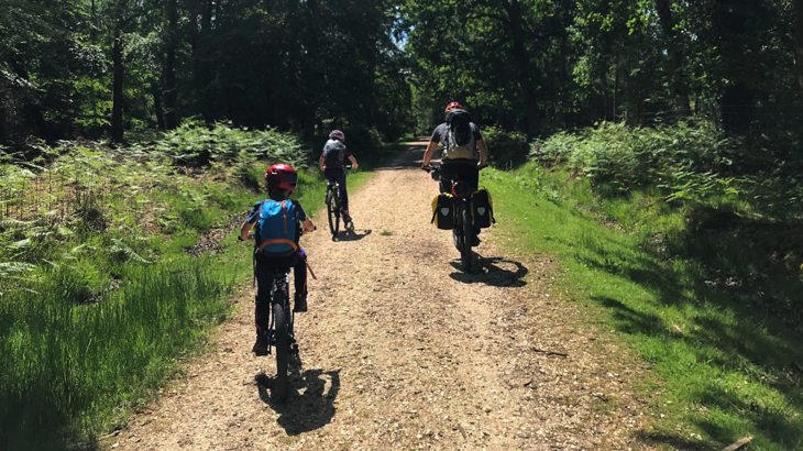 Father and two children cycling on gravel track through woodland. They are loaded with bags and panniers. It is a bright sunny day.