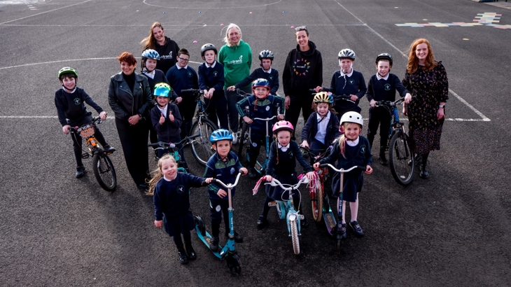 A group of children stand in a school playground with five female adults. Most of the children are on bikes or scooters and are wearing helmets.