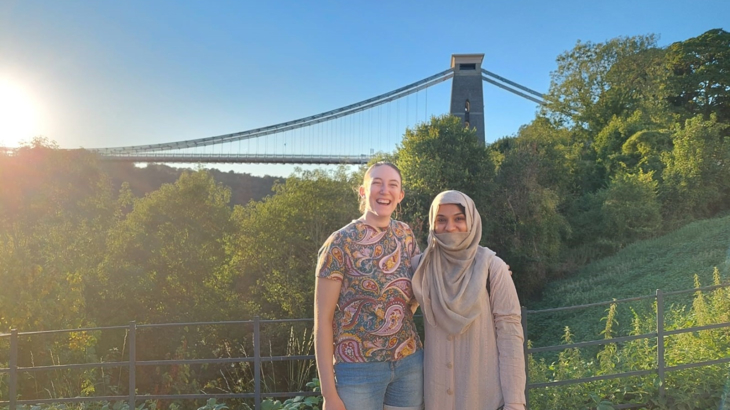 Two young women stood close together smiling in front of Clifton Suspension Bridge in Bristol on a summer's day, with blue skies above.