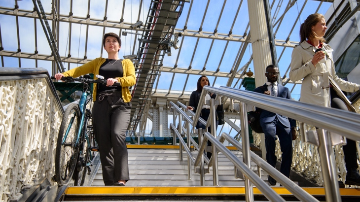 Four commuters in formal office wear walk down a staircase in a glass roofed railway station. One is wheeling a bike on a custom bike ramp which runs parallel to the stairs. The sky beyond the glass ceiling is bright and blue.