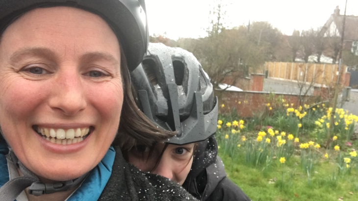 Kate Ball takes a selfie in the snow. She is wearing a cycle helmet and one of her children, also wearing a helmet, peeps over her shoulder.