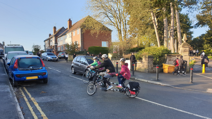 Kate cycles her electric tandem with her two children, one on the back, the other cycling beside. They are pulling onto a busy road on a steep incline, with oncoming traffic and parked cars.