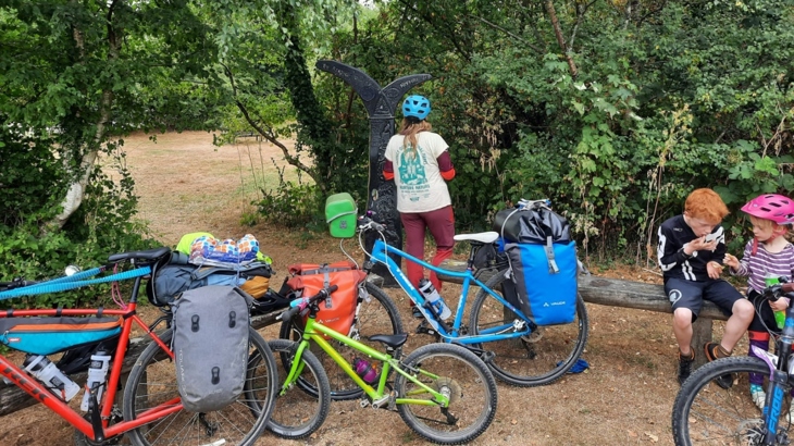 Emmer and her family are stopped on a traffic-free path on the National Cycle Network. Emmer is looking at a black painted Millennium mileposts, whilst her children are sat on a bench snacking. The family's bikes and luggage are all around them.