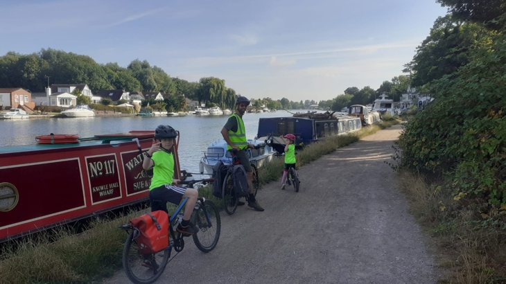 Emmer's husband and two children pose for a photo on the Great Western Way canal tow path. The three are stopped on their bikes beside narrow boats, the sky is blue and the sun is low in the sky.