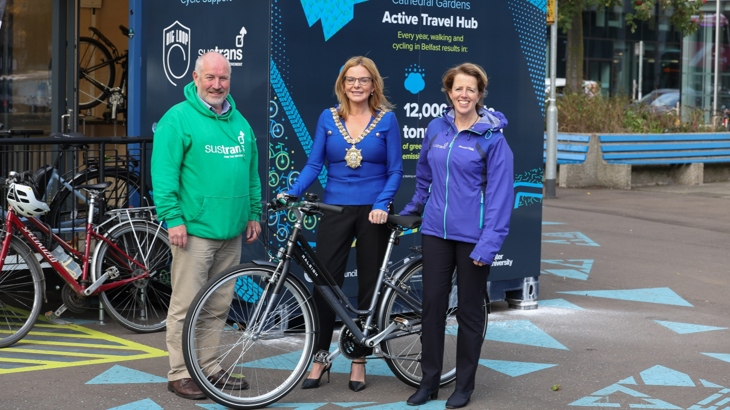 Belfast City Council Lord Mayor is stood holding a bike outside the new Cathedral Gardens active travel hub. Walk Wheel Cycle Trust colleagues are stood either side, all are smiling. Behind them are the blue graphics of the hub exterior, and the surrounding tarmac is painted with blue geometric patterns.