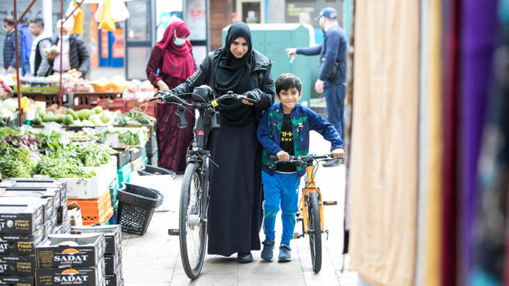 A child and and woman smiling while walking their bicycles through a busy fruit and vegetable market 