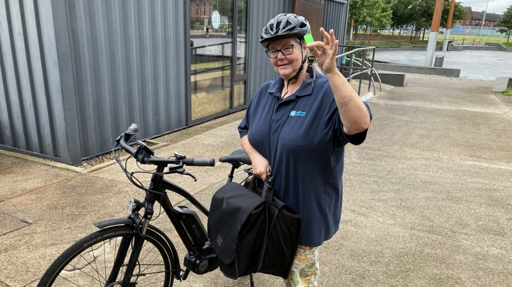 Sarah stands outside a cycle hub in Belfast with a black electric bike. She is wearing a helmet and smiling. The day looks dry and fine.