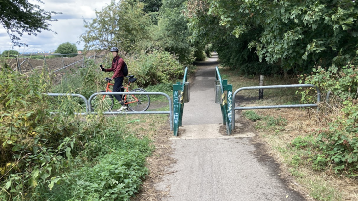 The photo shows a grey, gravelly path running through the middle, lined with overgrown shrubbery and trees. There is an A-frame type barrier across the path which is green with white graffiti. There is a person on the other side of the barrier on a red bike, facing towards the camera.