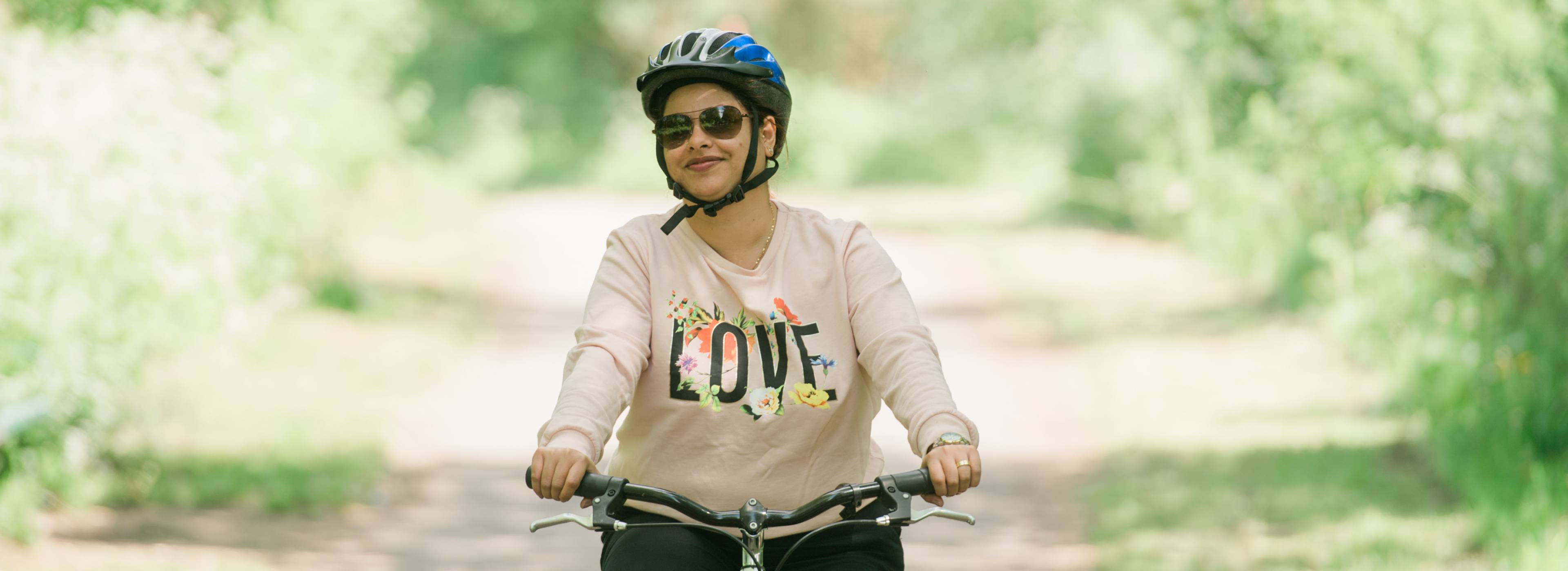 Smiling woman cycling through leafy green area on a sunny day. She is wearing a helmet and sunglasses.