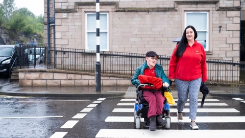 A man in a coat on a mobility scooter and a woman in a fleece, walking, are using a zebra crossing together. The road is wet and the day looks cool.