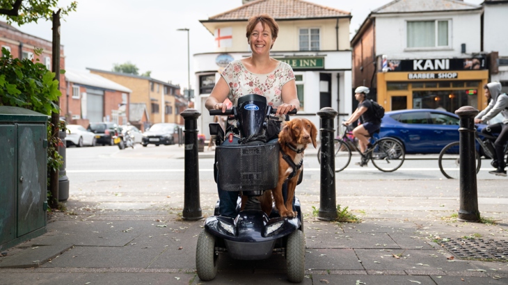 A smiling woman on a mobility scooter, with a dog at her feet. They are in a paved area with a shop-lined road in the background, beyond bollards. Some people are cycling in the background.