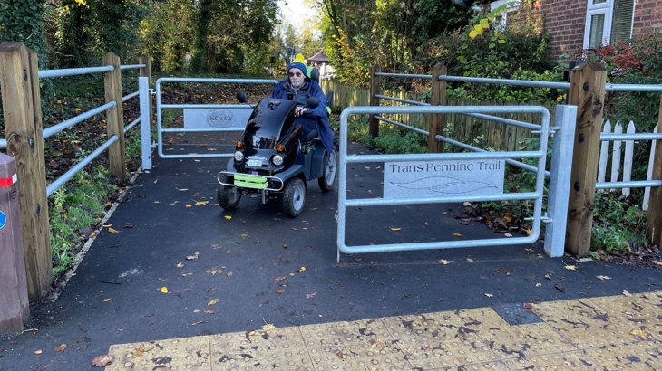 A man on a mobility scooter passes with ease through a new galvanised metal gate chicane on a traffic-free path on the Trans Pennine Trail. The chicane is wide and the path's surface looks newly tarmacked. In the foreground are raised tactile paving slabs.