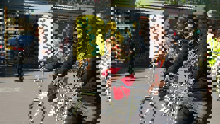 Woman with bicycle at a bike ride training course in a school playground with other adults cycling in background