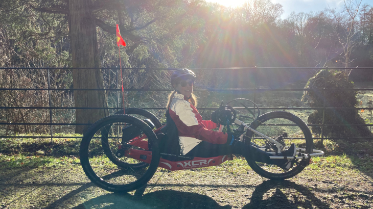 Laura sat smiling on her red XCR handcycle, on a traffic-free path. Behind Laura is countryside and trees. The sun is low in the sky.