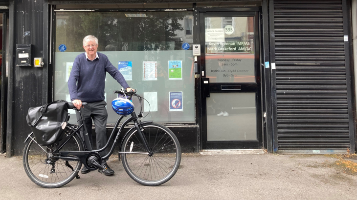 An image of Wales' First Minister, Mark Drakeford, smiling and looking into the camera whilst holding a black e-bike in front of his constituency office.
