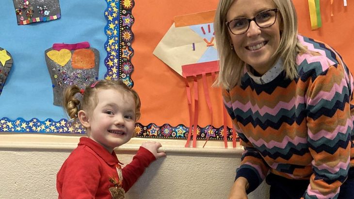 A little girl in a red jumper and a woman in a striped jumper are standing beside an indoor hall wall.