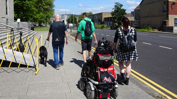 Four people, one with a guide dog and one in an electric wheelchair, walking together down a street and chatting
