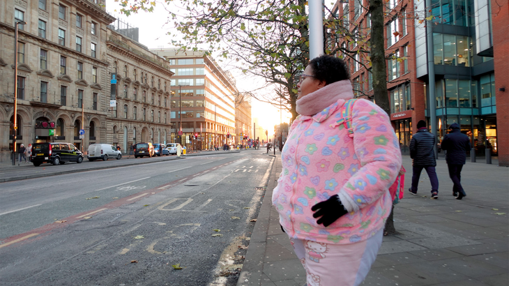 Alisha, standing on the pavement next to a main road waiting to cross, as the sun is setting behind her.