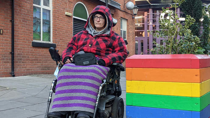 Dennis in her electric wheelchair, wheeling down a high street in Manchester past a rainbow-coloured planter box.