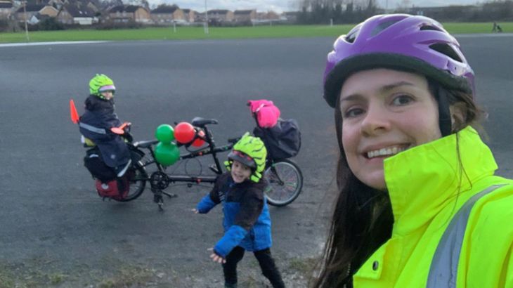 A woman wearing a purple helmet and hi-vis vest is outside taking a selfie photo of herself with her two small children and their cargo bike in the background.