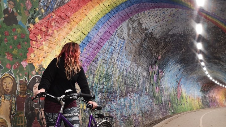 A woman with her bike walking through Colinton Tunnel in Edinburgh surrounded by a colourful mural all over the tunnels walls depicting a rainbow, people and nature scenes.