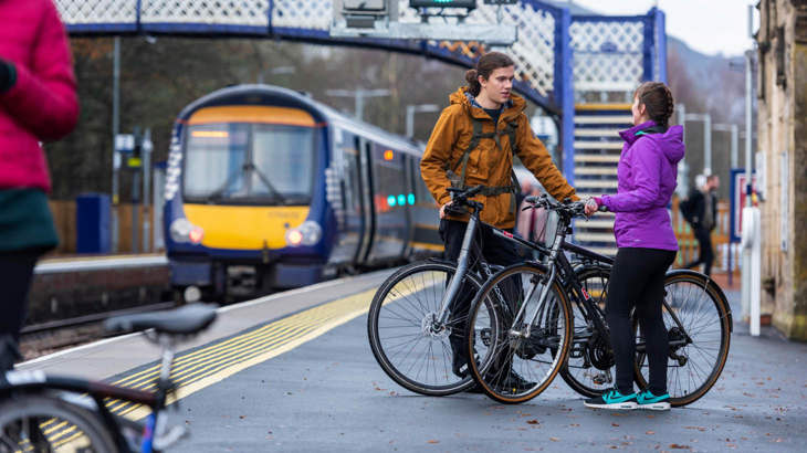 Two people with bikes standing at a train station as a train approaches the platform from the distance.