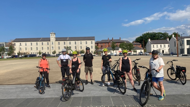 A group of women and men stand on bikes in an outdoors square with a bright blue sky.