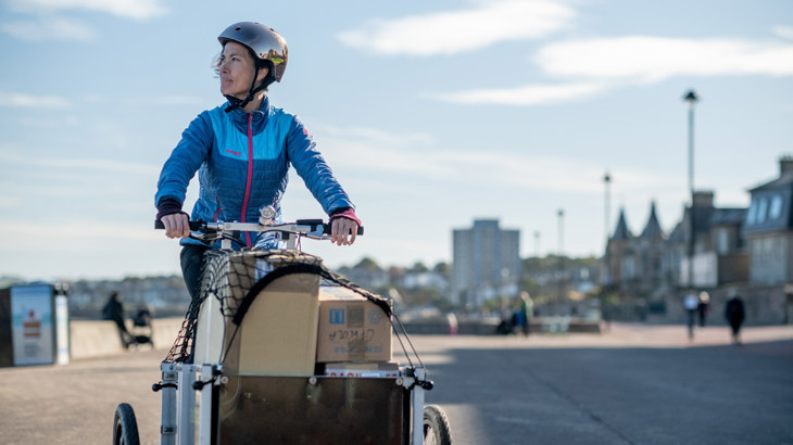 A woman rides a cargo cycle laden with boxes against the backdrop of a clear sky.