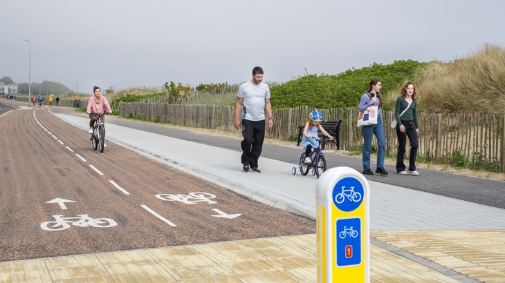 A cyclist approaches from the distance on a cycle lane. A child is pictured on a bike, accompanied by an adult. Separately, two adults are walking.